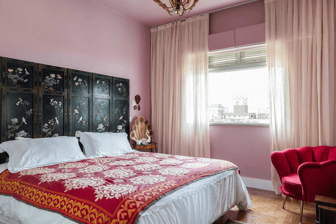 Quarto rosa com parede rosa, teto colorido rosa, biombo, roupa de cama branca, manta colorida estampada, cortina de verão, poltrona vermelha e piso de taco de madeira. 