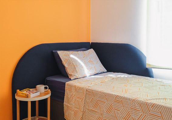 Vista angulada da área da cama do quarto. À esquerda, parede pintada na cor Polpa de Pêssego, e à direita, umaparede branca. A cama de solteiro está encostada nessas duas paredes, sendo a parte de cima na parede laranja e a lateral na parte branca. Sua cabeceira em veludo azul escuro está fixada em ambas as paredes, formando um L na parte de cima do colchão. O enxoval é azul escuro, branco e laranja e, à esquerda da cama, há uma mesa de apoio redonda com livros e uma xícara.