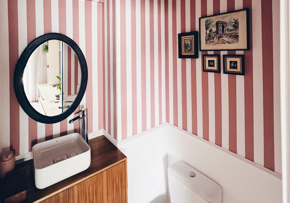 banheiro com parede decorada com listras rosa e branca, espelho oval com bordas pretas e pia com suporte de madeira representando tinta fosca ou acetinada