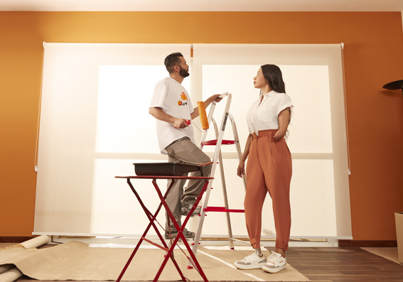 casal ao lado de parede branca com homem subindo na escada com rolo de pintura ao lado de banco com bandeja de tinta