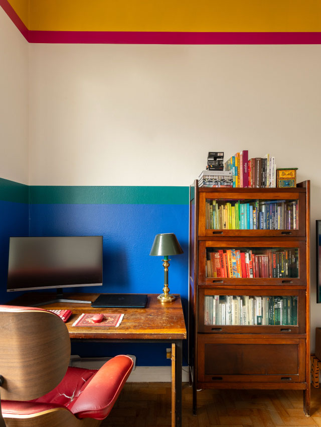 Imagem de um escritório com paredes verde, azul e branco em frente a uma mesa e estante com livros